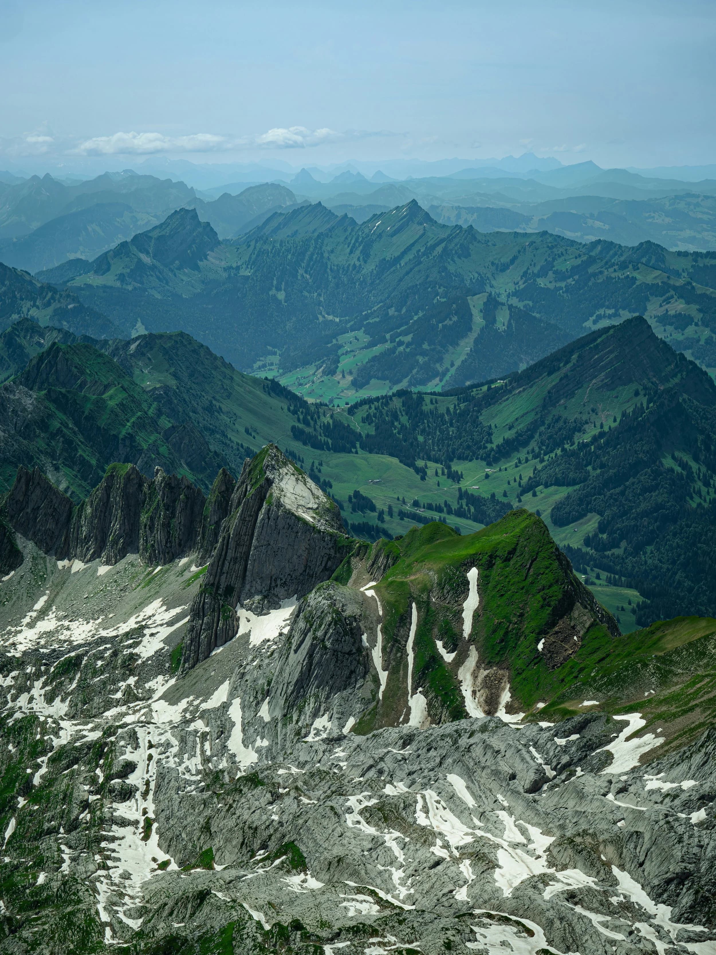 A mountain landscape with rugged peaks and patches of snow, green slopes, and a distant range of mountains under a cloudy sky.